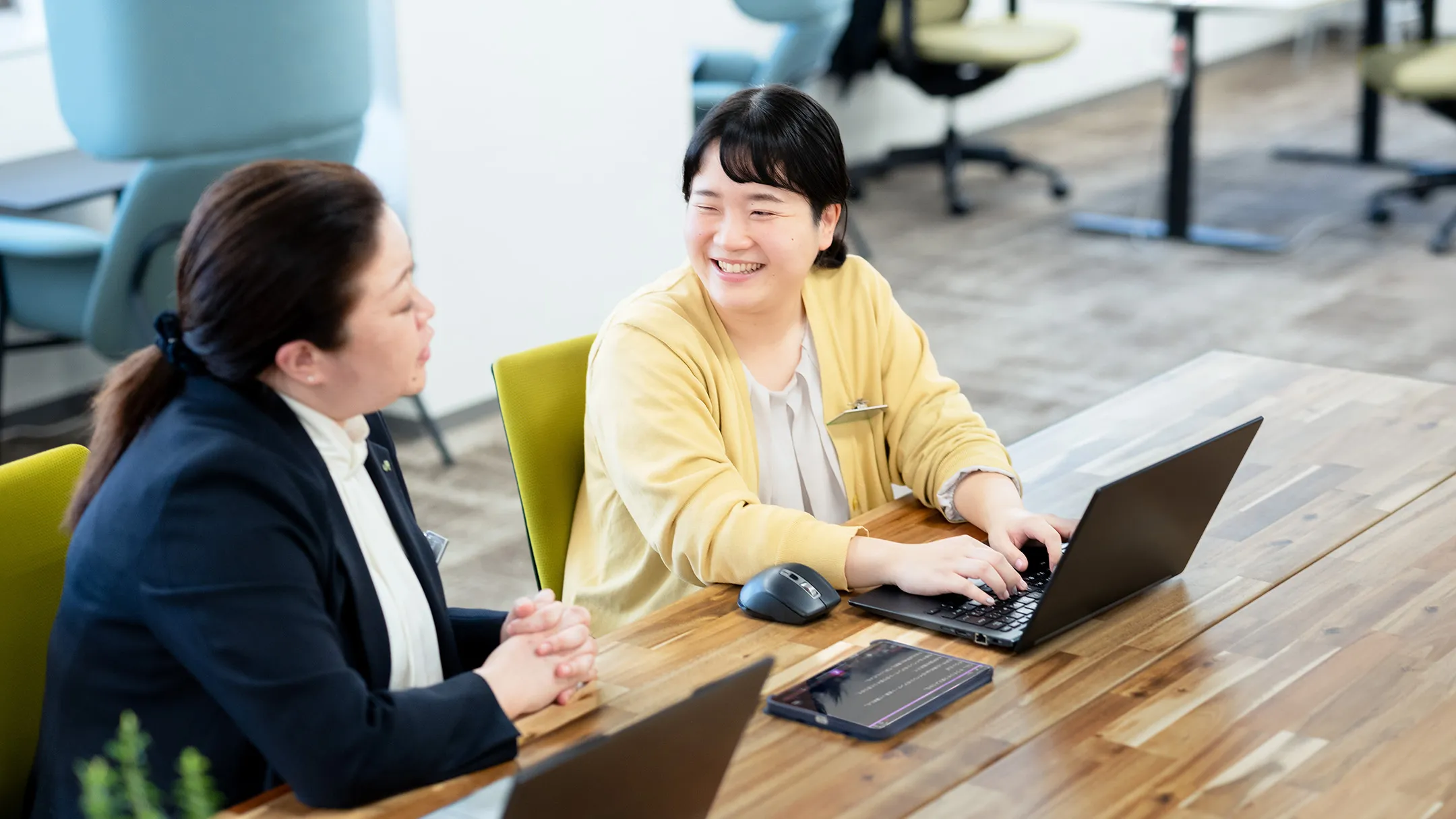 オフィスのデスクで、音声認識アプリを起動したタブレットを使い、キーボードを打ちながら同僚と笑顔で会話をする社員の写真